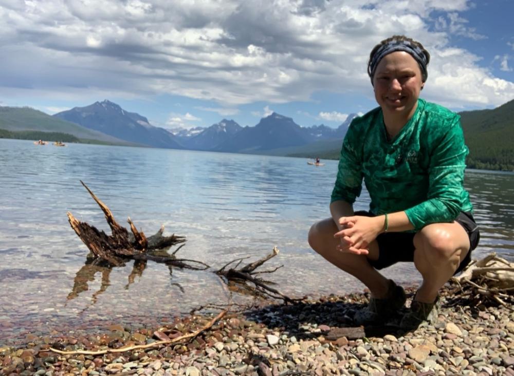 Breana is crouching on a rocky beach in front of a lake and mountains in the distance. She is smiling, wearing a bandana, a blue-green long t-shirt with the sleeves rolled up, shorts, and sneakers.