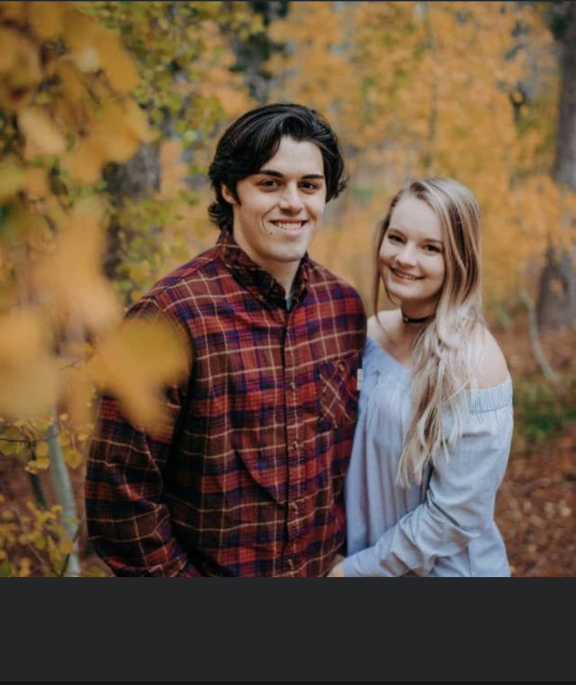 Logan is standing with his wife with fall woods in the background. Logan is smiling, has short brown hair, and is wearing a red plaid shirt. His wife is smiling, has long blonde hair, and is wearing a black choker and white off the shoulder shirt.