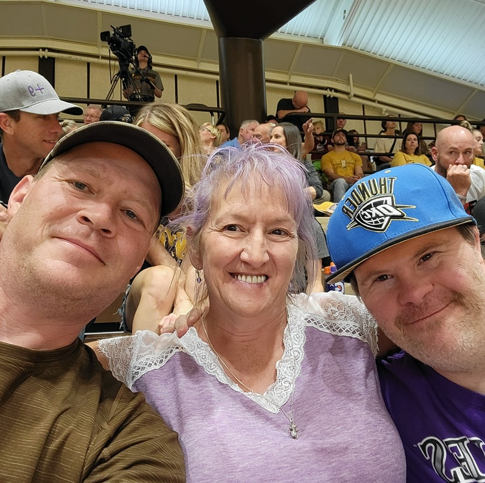 Michelle is in the middle of a group of three with her arms around the others, and in the bleachers of a UW game. She is smiling, has purple hair that is tied up, and is wearing a dainty necklace and purple shirt with white lace details. On her left is her husband Brian, who is smiling and wearing a brown shirt and baseball cap. On her right is a (male) participant who is smiling and wearing a Rockies shirt and Thunder snapback.