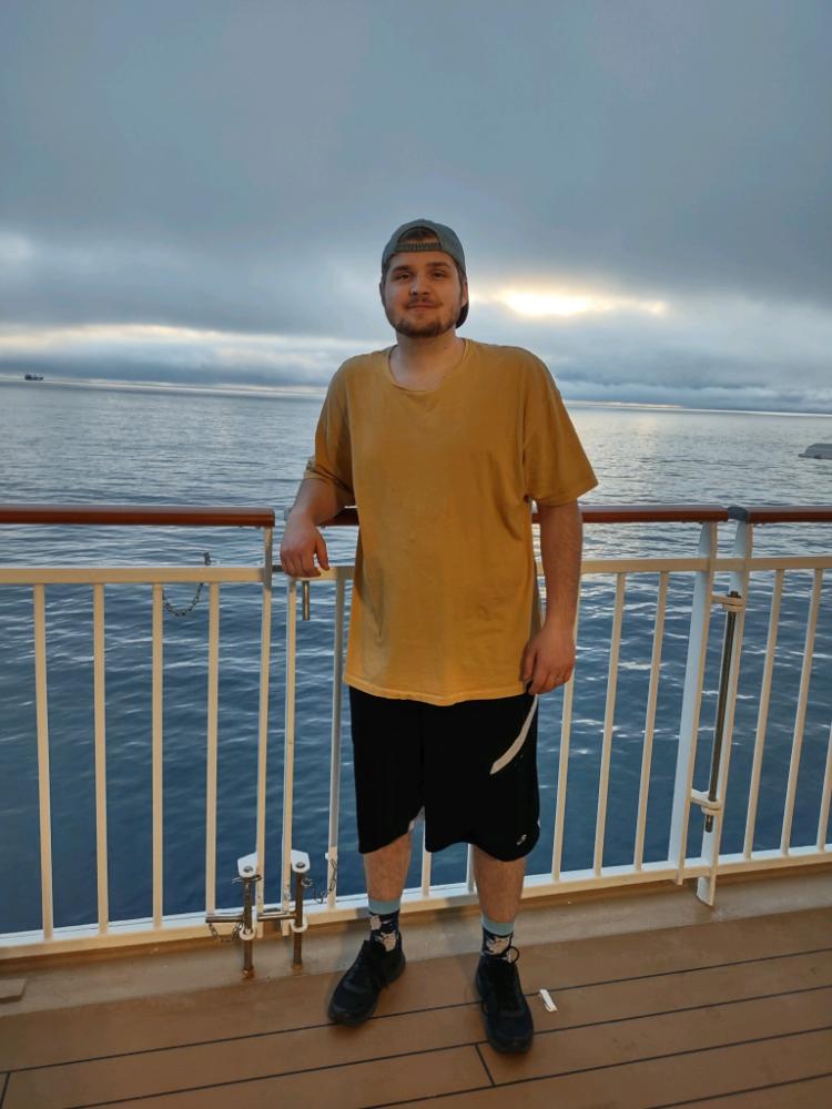 Mondell is standing in front of railing with the ocean in the background. He's smiling and wearing a yellow shirt and backwards cap.