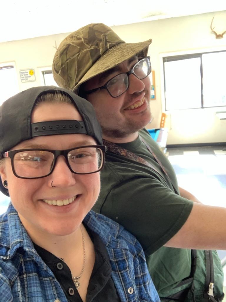 Two people are smiling for a selfie in a well-light room, but the one on the rights' torso is facing to the right and they're almost behind the other person. He is also wearing glasses, a dark t-shirt, a fanny pack and a fishing hat. The person on the left is wearing a backwards ball cap and glasses. She is also wearing a blue flannel over a black shirt and a thin necklace.