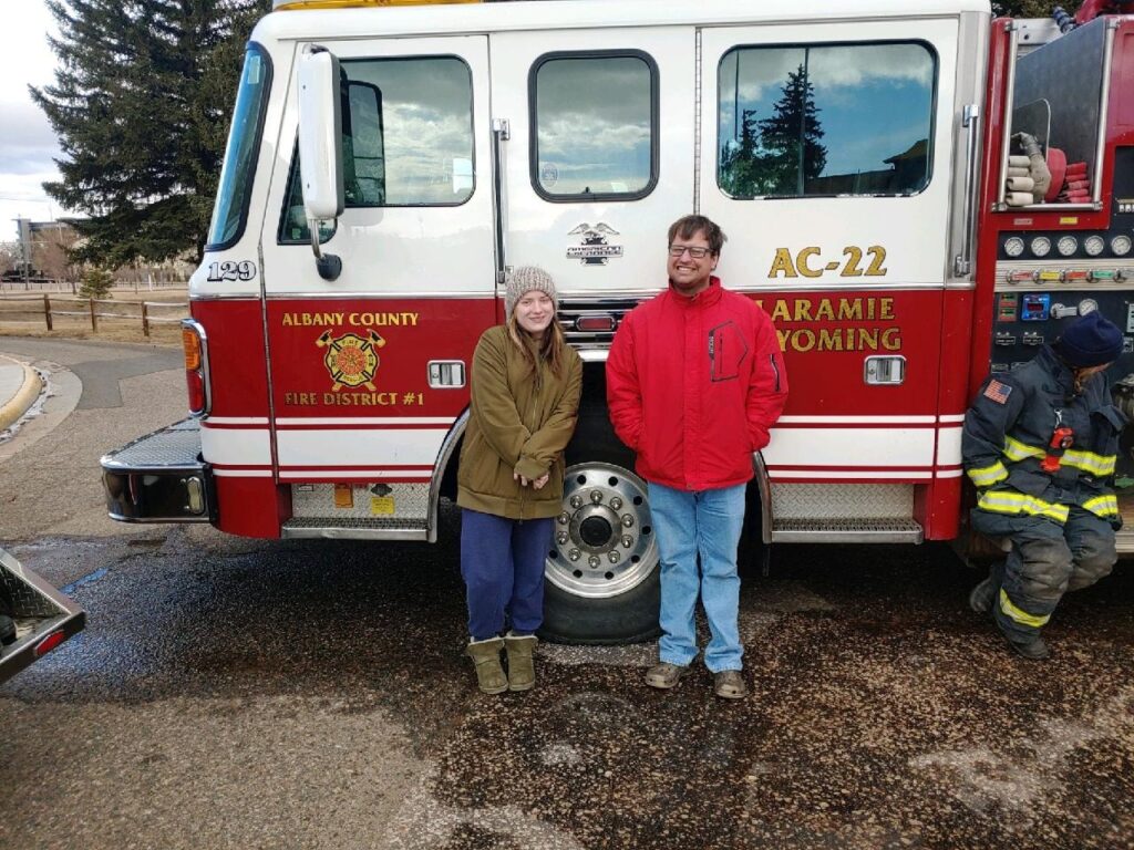 Two people are standing in front of a fire truck smiling for the photo. On the left, she is wearing a grey beanie, brown coat, blue pants and Uggs. On the right, he is wearing a red coat, jeans and Crocs. On the far right there is an additional fire fighter sitting and looking away.