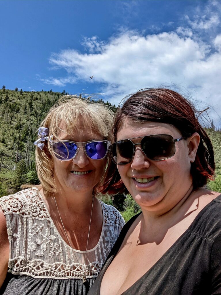 Two people are smiling for a selfie with a background of a forested mountain and sky. On the left, she is wearing dark sunglasses with a dark shirt and her dark brown hair is in a short bob. On the left, she is wearing a patterned white shirt and blue/purple colored sunglasses, with her light hair in a short bob with bangs.