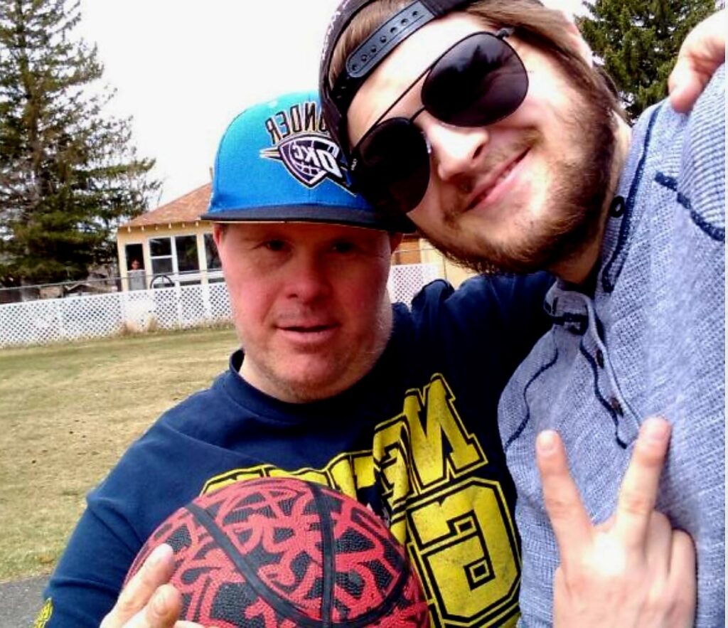 Two people are standing in a park taking a selfie in the summer. The one on the right is holding up a peace sign. He is wearing sunglasses, a backwards cap, and a grey sweatshirt. The one on the left is holding a black and red basketball. He is wearing a blue snap back with a team logo and a black t-shirt with yellow words that are mostly covered.