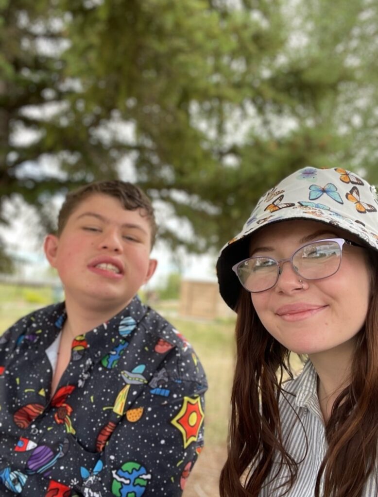 Two people are smiling and taking a selfie in front of a tree in the summer. On the left is a male with short brown hair and a colorful button down shirt. On the right is a female with long brown hair, glasses, and a bucket hat with butterflies.