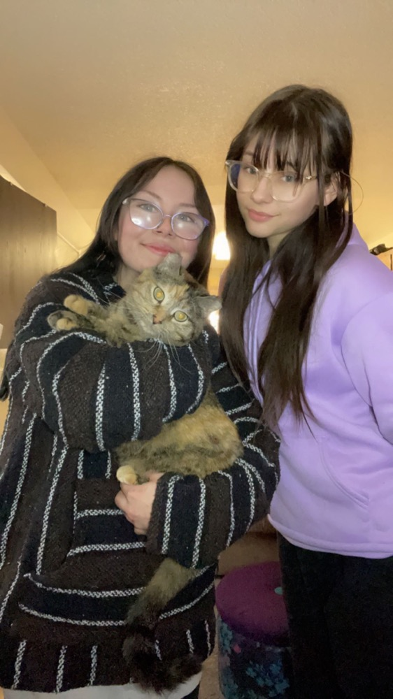 Zada is standing on the left with her sister next to her and holding her cat. She is smiling, wearing glasses and a patterned sweater with her brown hair down. Her cat is a patterned tabby who is staring straight into the camera. Her sister is also smiling, wearing glasses, and has her brown hair down. She is wearing a purple sweater.